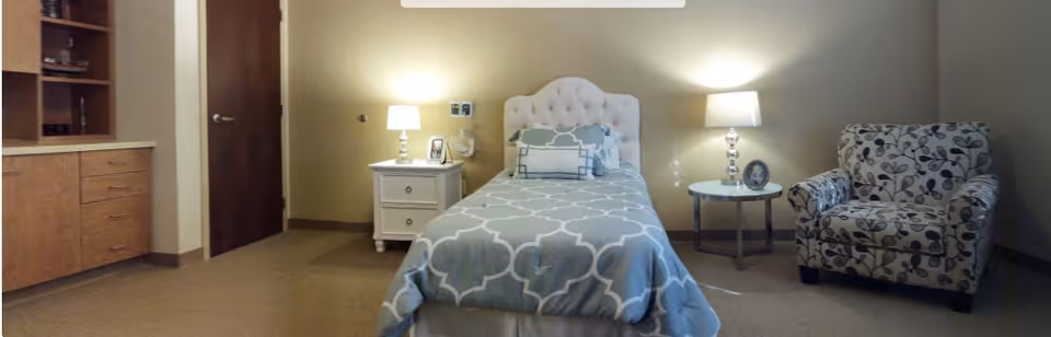 A softly lit assisted-living bedroom with a single bed, two bedside tables and lamps, a patterned armchair, and built-in cabinetry.