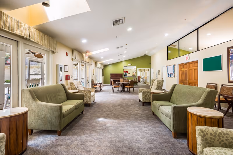 A spacious and well-lit common area in a senior living facility with green sofas, patterned armchairs, round wooden side tables, and a carpeted floor. The room has large windows with valances on the left side, letting in natural light. At the far end, there is a piano and a table with chairs. The walls are painted in light colors with one green accent wall, and there are bulletin boards and framed pictures on the right wall.