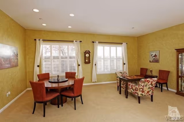 A well-lit room with two sets of tables and chairs. On the left, a round wooden table with four red upholstered chairs. On the right, a rectangular wooden table with two floral upholstered chairs and two red upholstered chairs. The walls are painted yellow with two large windows covered by white curtains and blinds. A wall clock hangs between the windows, and there is a glass display cabinet on the right side of the room. A painting is hung on the left wall.