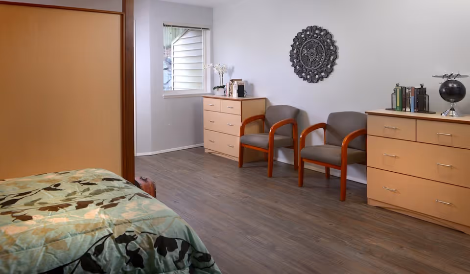 A bright furnished bedroom with a partially visible bed, two wooden-framed chairs, and dressers against a wall with decorative wall art.