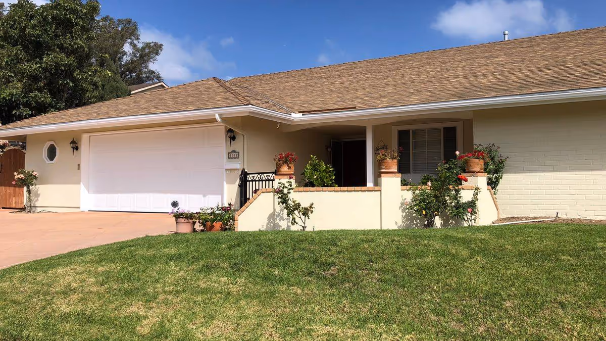 Front exterior of a single-story house with an attached garage, small porch with potted plants, and a green lawn.