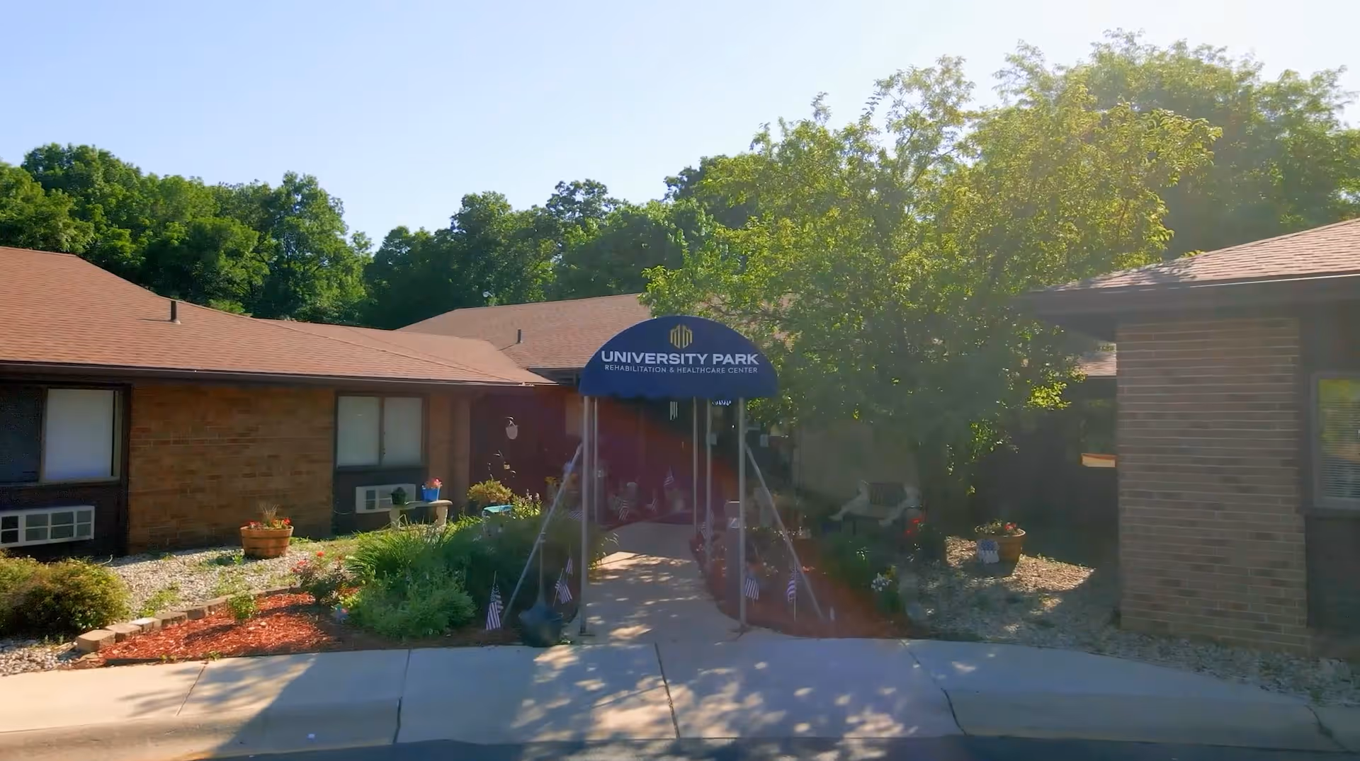 Exterior view of University Park Rehabilitation and Healthcare Center showing a single-story brick building with a covered entrance. The entrance has a blue canopy with the facility's name on it. There are plants, small American flags, and trees around the entrance area under a clear sky.