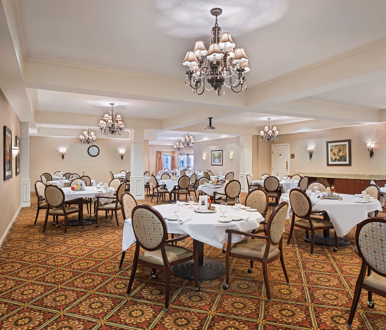 A spacious dining room with multiple round tables covered with white tablecloths, each set with glassware, plates, and napkins. The room features patterned carpet flooring, elegant chandeliers hanging from the ceiling, wall sconces, framed artwork, and a clock on the far wall. The overall ambiance is warm and inviting.