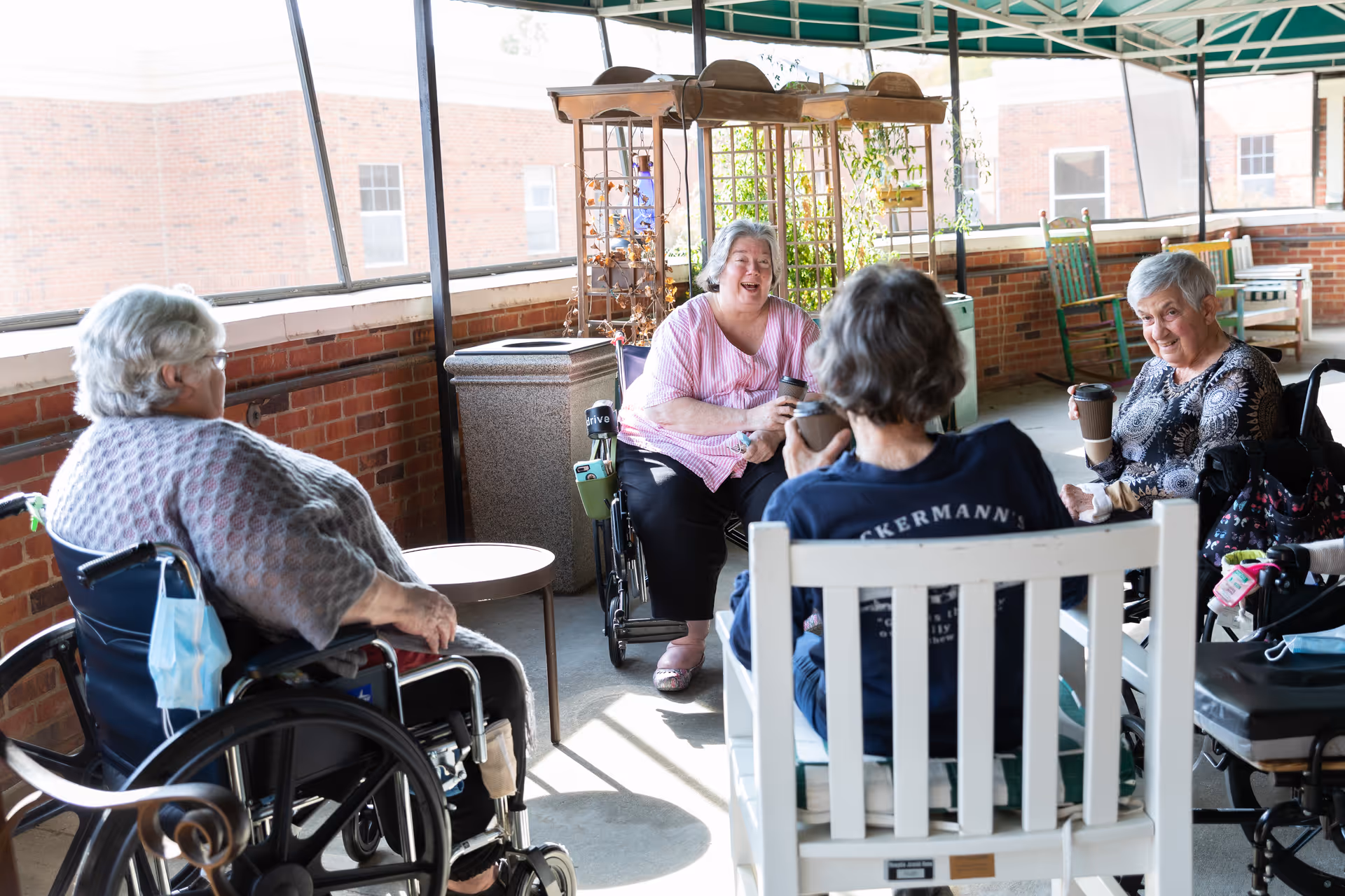 Four elderly women sitting and chatting on a covered outdoor patio area. Two women are in wheelchairs, one woman is sitting on a white wooden chair, and another woman is seated in a regular chair. They are holding coffee cups and appear to be enjoying a social moment together. The patio has brick walls, potted plants, and rocking chairs in the background.