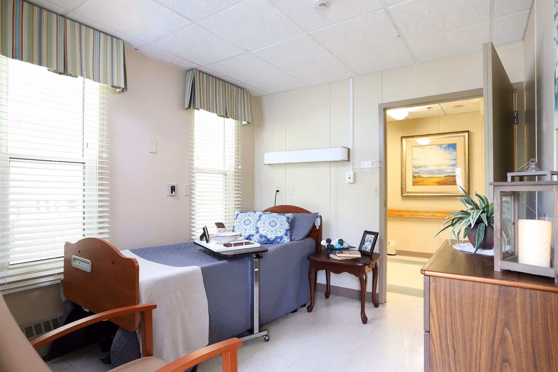 A bright senior living bedroom with a single medical-style bed, bedside table, dresser with decor, chair, large windows, and an open door to a hallway.
