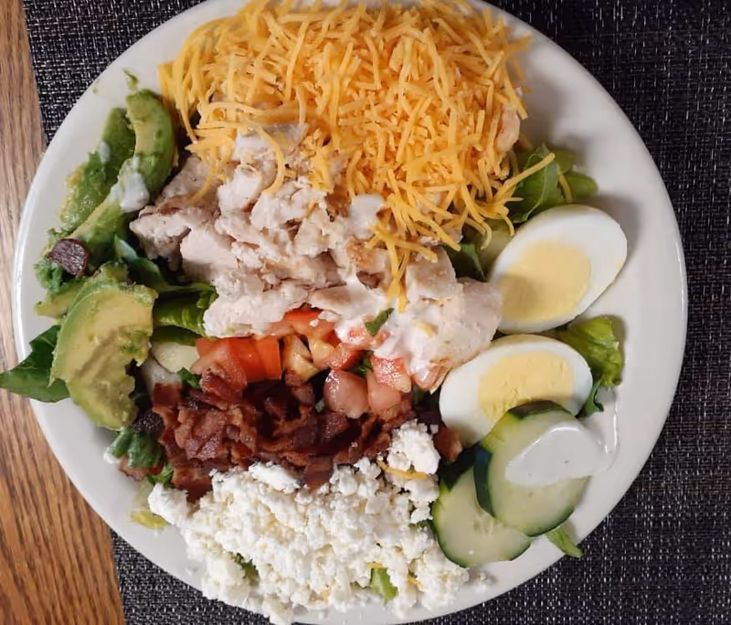 A plate of salad with shredded cheddar cheese, grilled chicken pieces, sliced avocado, chopped tomatoes, bacon bits, crumbled cheese, cucumber slices, and two halves of a hard-boiled egg, all on a bed of lettuce with a drizzle of dressing.