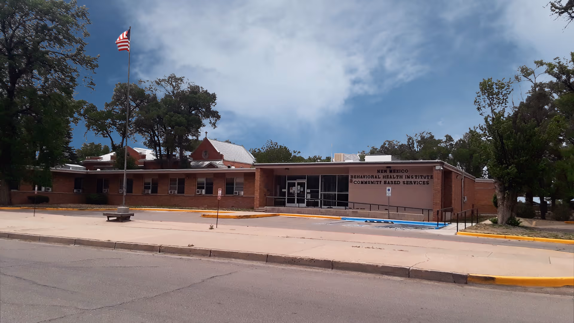 Exterior view of the New Mexico Behavioral Health Institute Community Based Services building, a single-story brick structure with a flat roof, an American flag on a flagpole in front, and trees surrounding the area under a partly cloudy sky.
