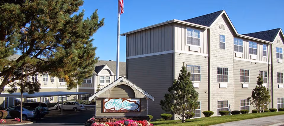 Front exterior of a multi-story senior living building with a Flagstone sign, landscaped flowerbeds, covered parking and an American flagpole.