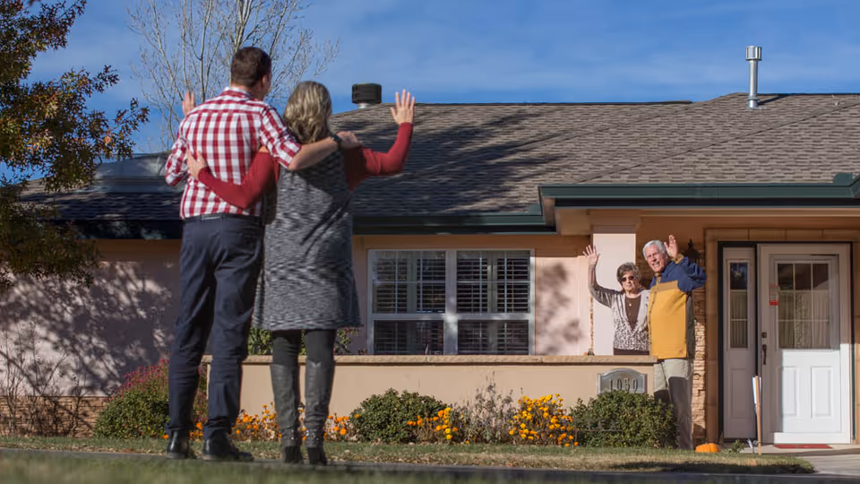 Two people standing outside with their backs to the camera, waving towards an elderly couple who are standing on the porch of a single-story house, also waving back. The house has a beige exterior with a white door and windows, and there are flowers and shrubs in front of the porch.