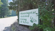 A white sign with green text reading 'Morningstar Assisted Living of Sylva' sits beside a tree-lined road.