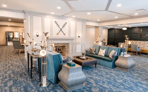 A spacious and well-lit living room area with blue patterned carpet, teal sofas with decorative pillows, a brick fireplace with crossed oars mounted above it, modern floor lamps, and a seating area with tables and chairs in the background.