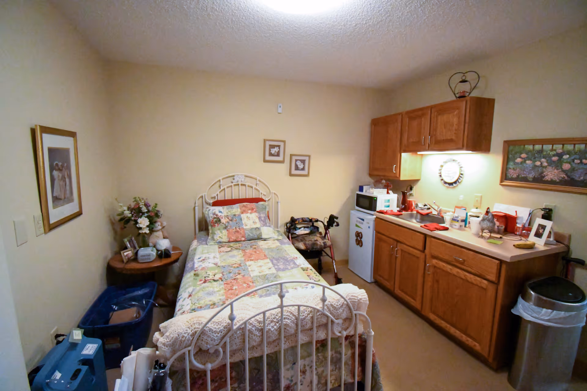 A small senior living facility room featuring a single bed with a floral patchwork quilt and a crocheted blanket at the foot. To the right of the bed is a kitchenette area with wooden cabinets, a microwave, a mini fridge, and various items on the countertop. On the left side of the bed is a round wooden side table with a flower arrangement, a teddy bear, and framed photos. The walls are beige and decorated with framed pictures. A trash can is visible near the kitchenette.