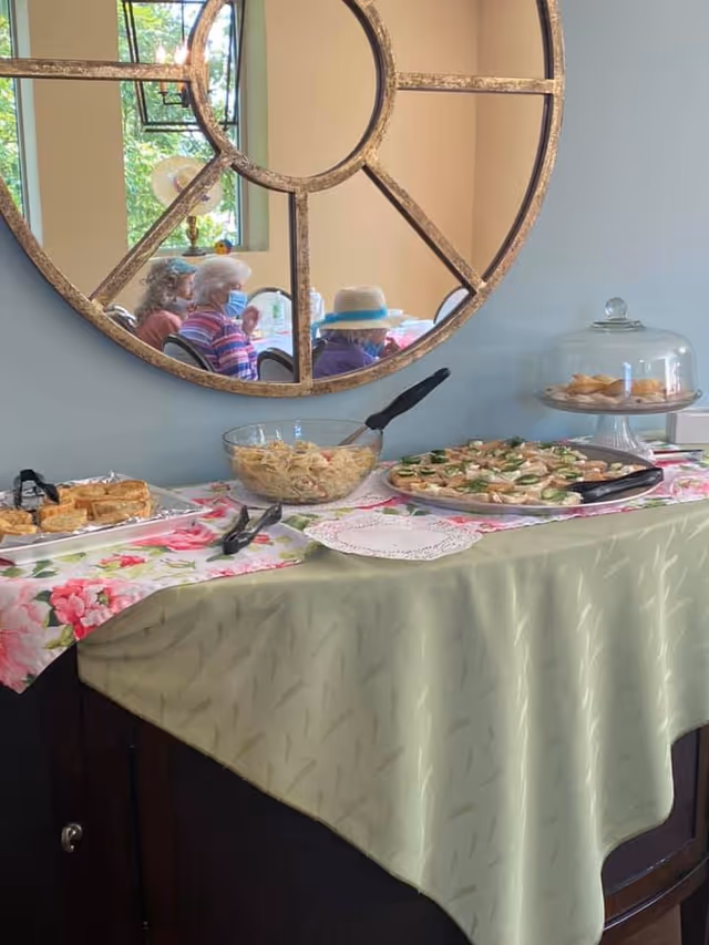 A buffet table covered with a green tablecloth and a floral runner, displaying plates of appetizers, a bowl of pasta salad, and a glass cake stand with pastries. A large decorative round mirror on the wall reflects three elderly people sitting at a table, two wearing masks and one wearing a hat.