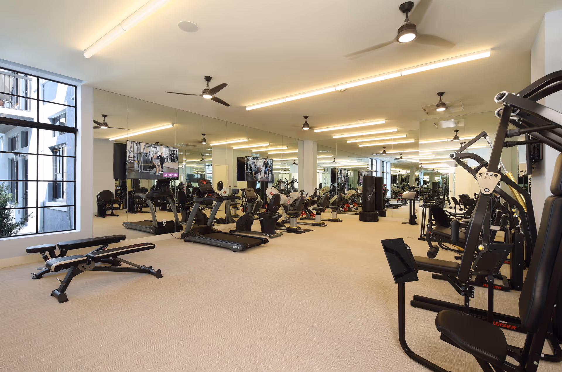 A spacious and well-lit fitness room with various exercise equipment including treadmills, stationary bikes, weight machines, and benches. Large mirrors cover one wall, reflecting the equipment and ceiling fans. A large window allows natural light to enter the room.