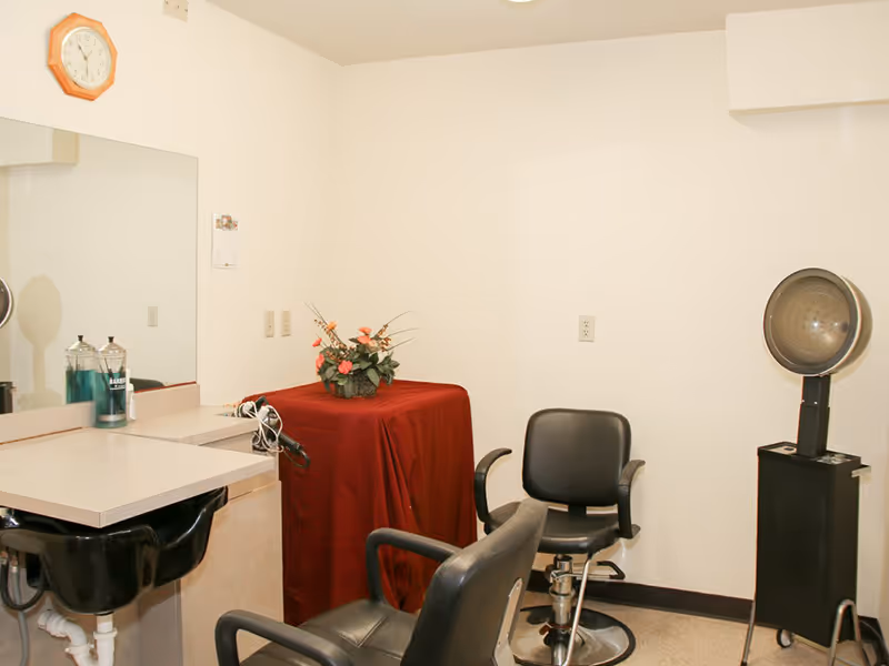 Interior of a small salon or hair styling area with two black salon chairs, a black hair washing sink, a large mirror, a clock on the wall, a covered table with a flower arrangement, and a vintage hair dryer.