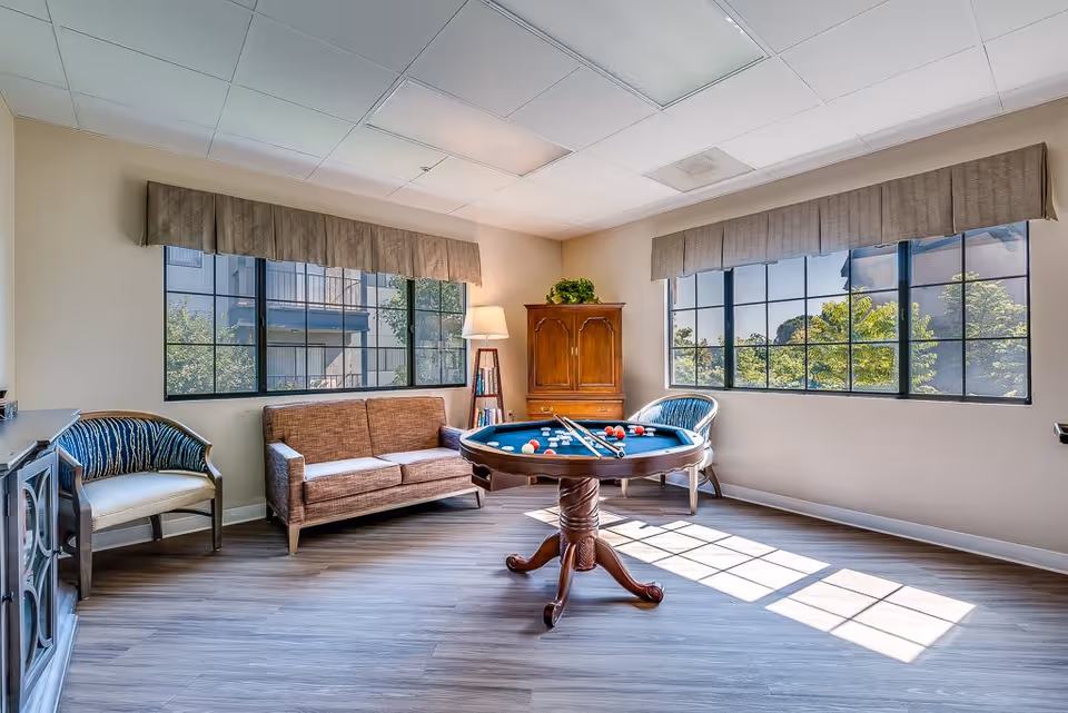 A bright room with large windows letting in natural light, featuring a round pool table with billiard balls and cues, a brown sofa, two armchairs with blue and white upholstery, a wooden cabinet with a plant on top, and a floor lamp next to a small bookshelf.