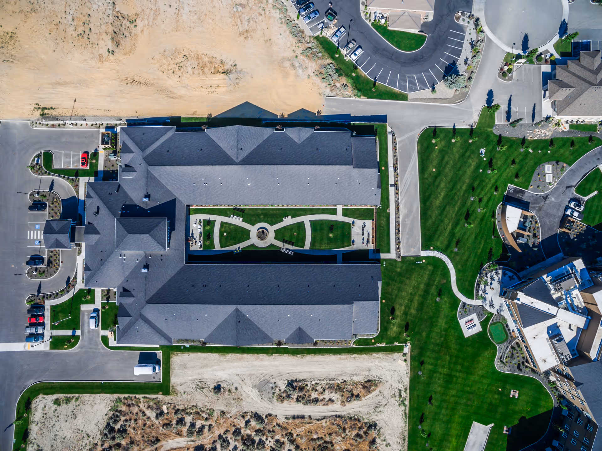 Aerial view of a large building complex with a central courtyard featuring a circular pathway and green lawn. Surrounding the building are parking lots, roads, and landscaped grassy areas with trees and pathways.