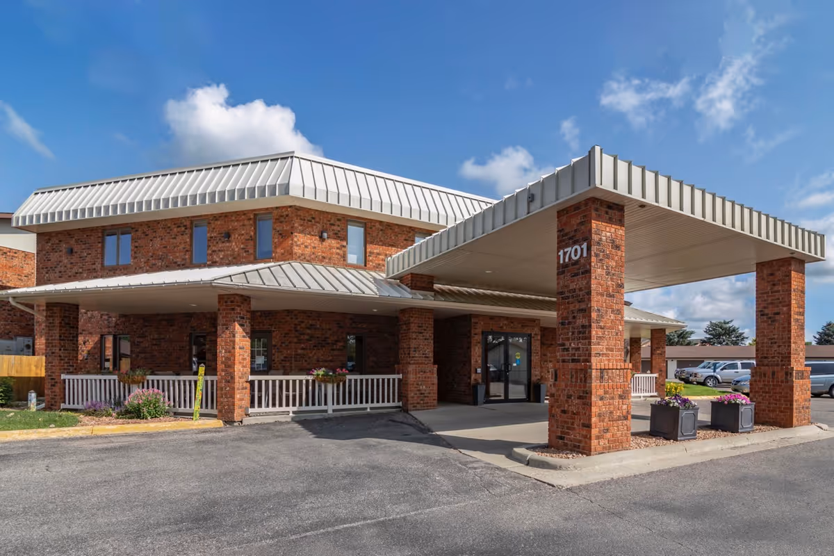 Exterior view of a senior living facility building with red brick walls and a white metal roof. The entrance has a covered drop-off area supported by brick pillars, with the number 1701 displayed on one pillar. There are flower pots with colorful flowers near the entrance and a parking lot with several vehicles in the background under a partly cloudy blue sky.