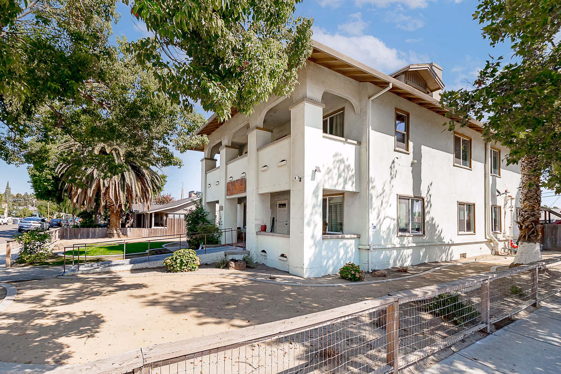 Exterior view of a two-story white building with a covered balcony and several windows, surrounded by trees and a fenced yard under a clear blue sky.