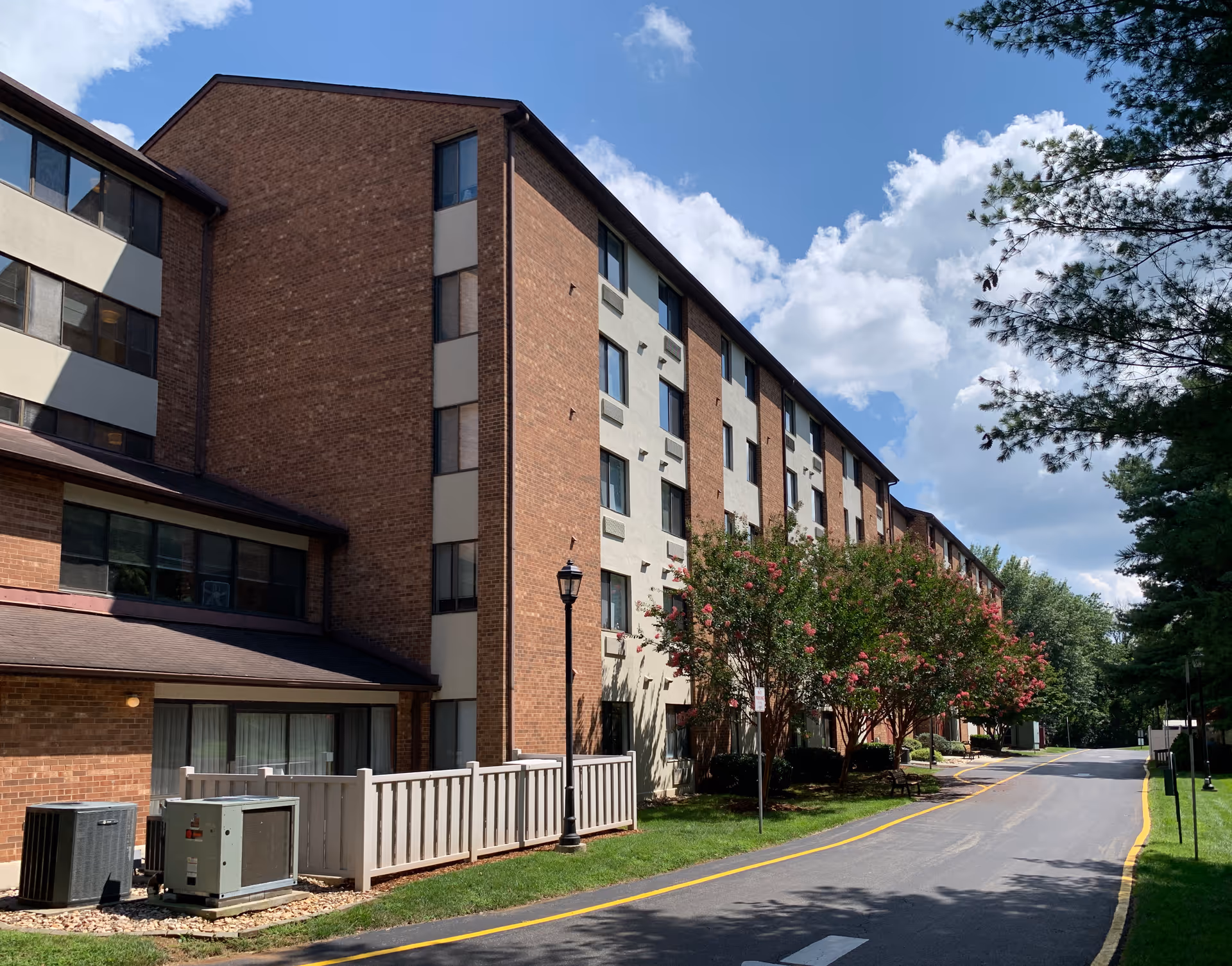 Exterior view of a multi-story brick and beige building with several windows, a paved road with yellow lines running alongside it, green grass, flowering trees, and a clear blue sky with some clouds.