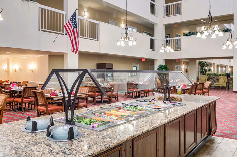 Interior view of a dining area in Dunwoody Pines Retirement Community featuring a salad bar with various fresh vegetables and toppings under a glass sneeze guard. The room has multiple dining tables with chairs, red patterned carpet, chandeliers hanging from the ceiling, and an American flag mounted on a pillar. The space is open with a second-floor balcony overlooking the dining area.