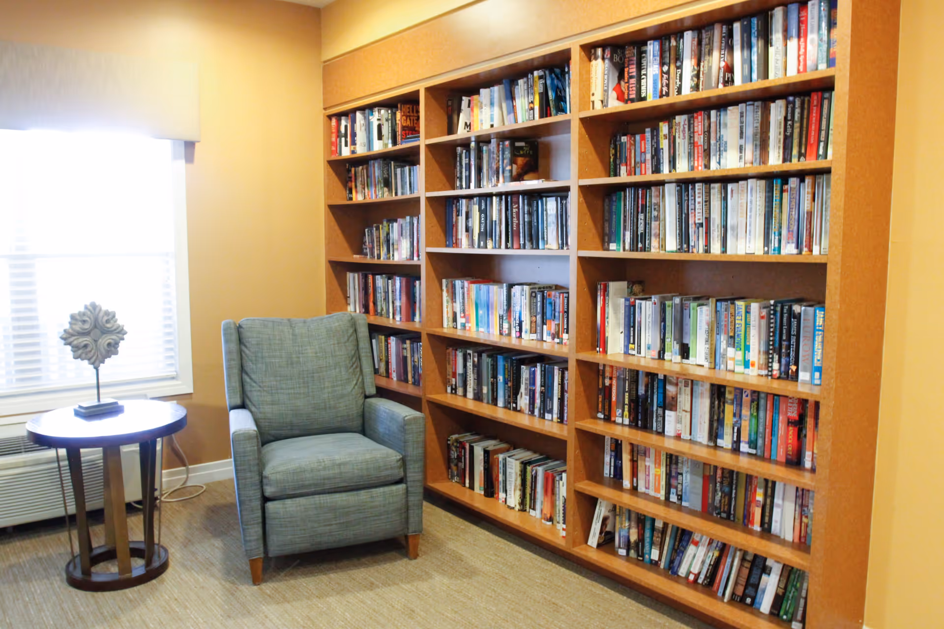 A cozy reading nook featuring a light blue upholstered armchair next to a round wooden side table with a decorative sculpture. Behind the chair is a large wooden bookshelf filled with numerous books. A window with white blinds allows natural light into the room.