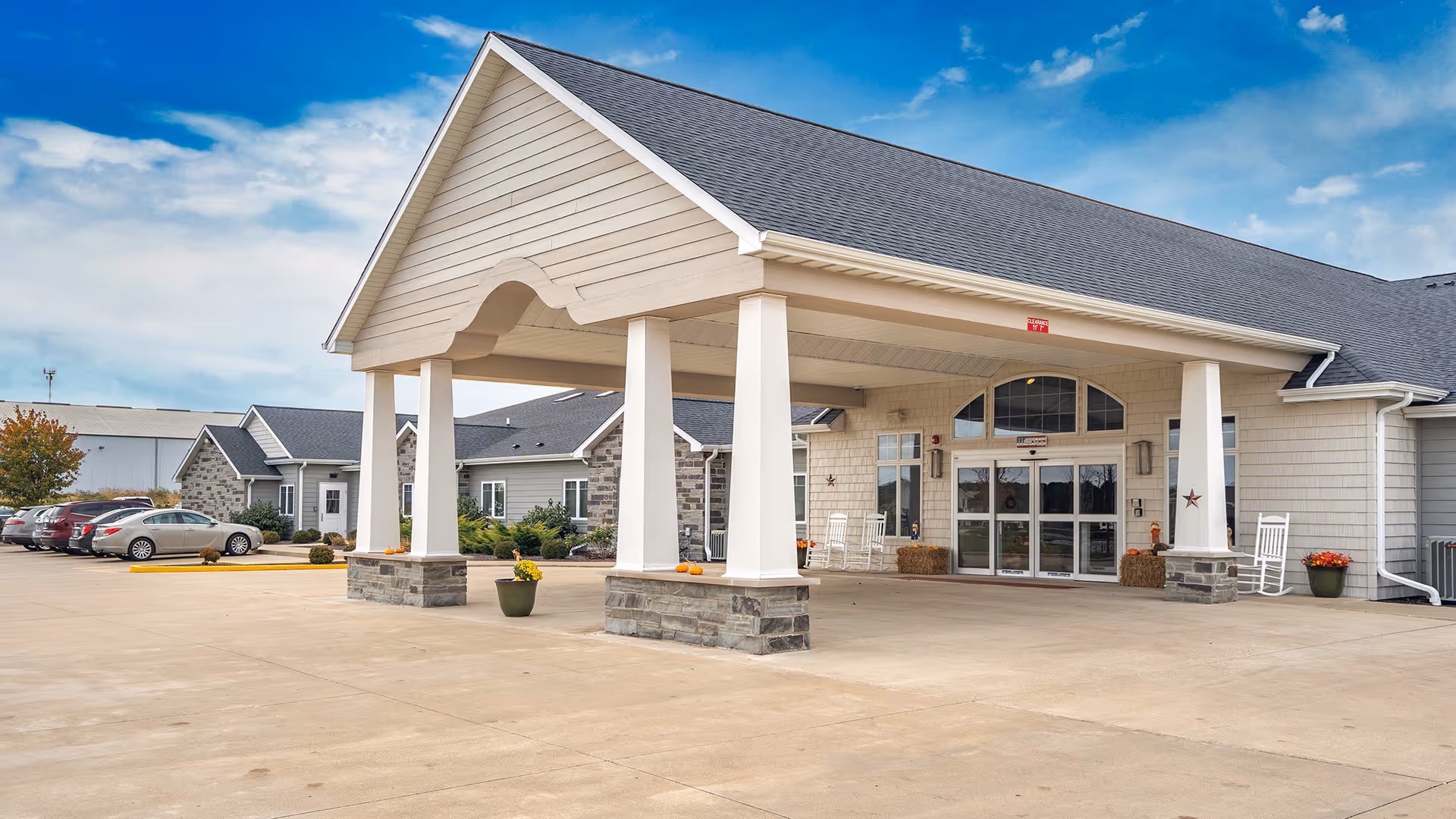 Exterior view of Reflections Memory Care facility in Washington, IL, showing the main entrance with a covered drop-off area supported by white columns with stone bases. Several cars are parked to the left, and the building features light-colored siding and a gray roof under a partly cloudy blue sky.