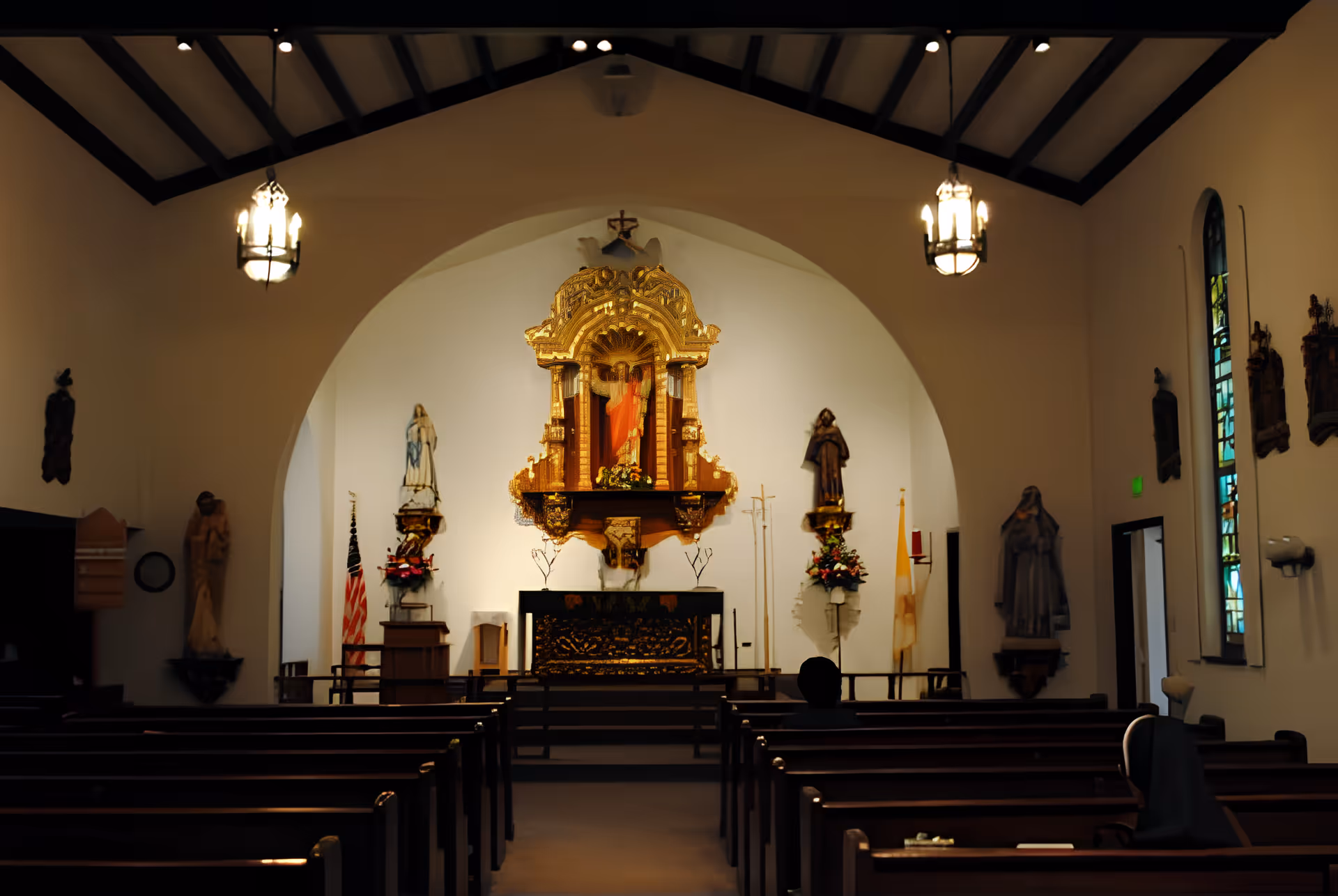 Interior view of a chapel with wooden pews facing an ornate altar, statues, and hanging lights.