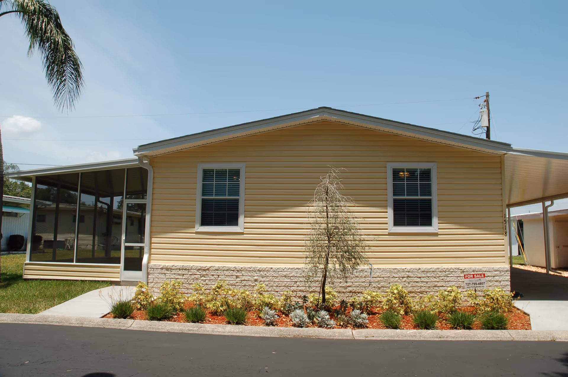 Front exterior of a yellow manufactured home with two windows, a screened porch on the left, a carport on the right, and a landscaped flowerbed with a 'For Sale' sign.