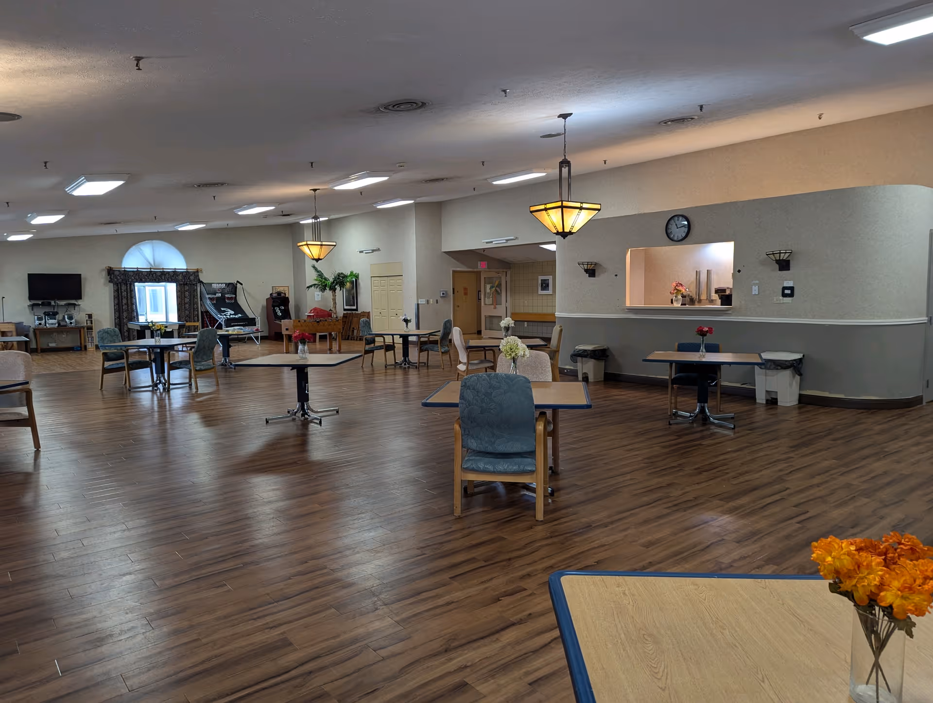 A spacious dining room with several tables and chairs arranged on a wooden floor. Each table has a small vase with flowers. The room has beige walls, ceiling lights, and hanging pendant lamps. There is a service window with a clock above it and a few trash bins nearby. In the background, there is a TV, a window with curtains, and some recreational equipment.