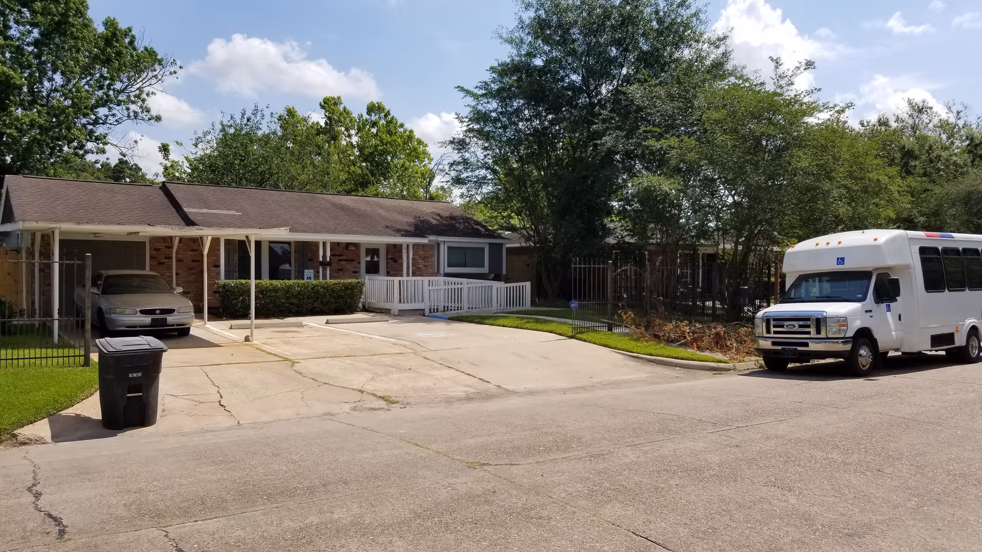 Exterior view of a single-story brick building with a covered carport and a white ramp leading to the entrance. A white shuttle bus is parked on the street in front of the building, and there is a black trash bin near the driveway. Trees and greenery surround the area under a partly cloudy sky.