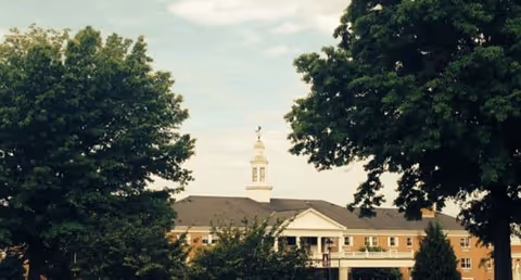 Exterior view of a multi-story brick building with a white cupola on the roof, partially obscured by large green trees in the foreground under a partly cloudy sky.