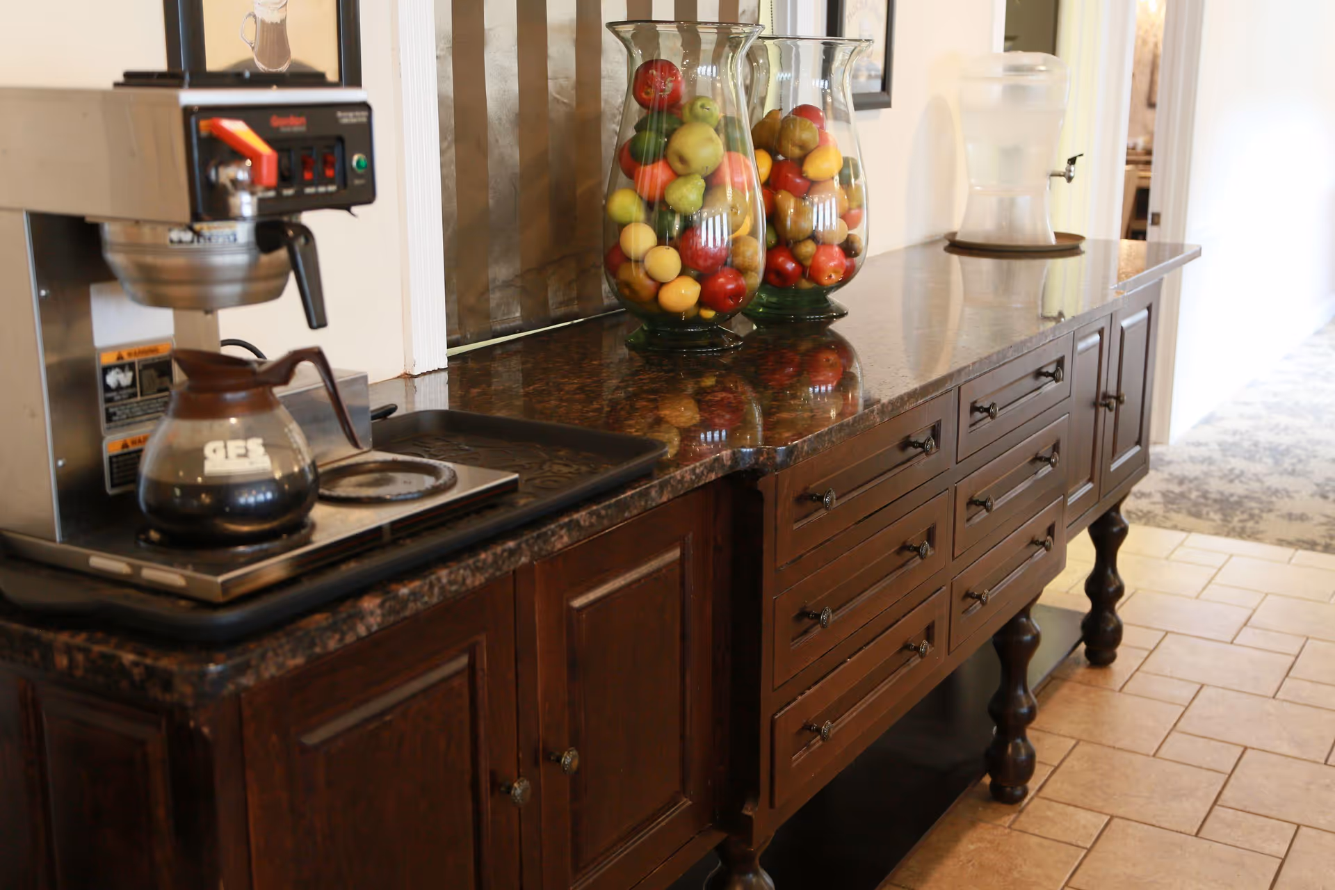 A dark wooden sideboard with a granite countertop holding a coffee maker with a glass pot of coffee, two large glass vases filled with artificial fruit, and a water dispenser. The sideboard is placed in a hallway with tiled flooring and a carpeted area visible in the background.