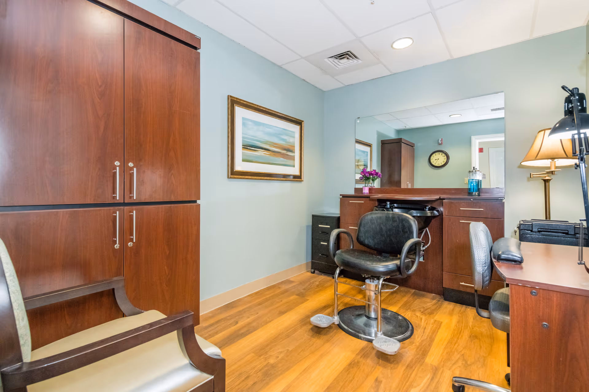 Interior view of a salon area in a senior living facility with a black salon chair in front of a large mirror, wooden cabinets, a small sink, a desk with a lamp, and a framed landscape painting on the wall.
