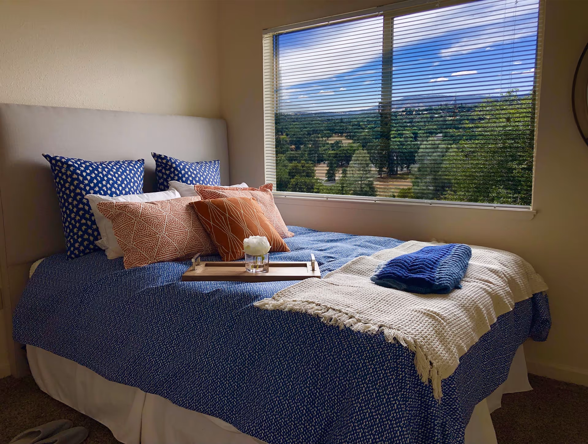 A neatly made bed with a blue bedspread, multiple decorative pillows in blue and orange patterns, and a white throw blanket with a folded blue towel on top. A wooden tray with a small white flower in a glass vase is placed on the bed. The room has a large window with blinds partially open, showing a scenic view of trees and a blue sky with clouds outside.