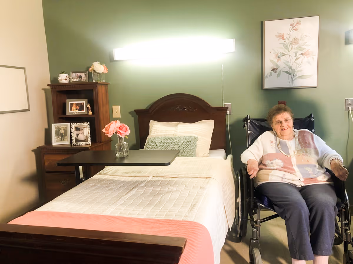 A senior woman sitting in a wheelchair next to a neatly made bed with a wooden headboard in a room with green walls. There is a small table with a vase of pink flowers on the bed, a wooden nightstand with framed photos and decorations, and a floral painting hanging on the wall.