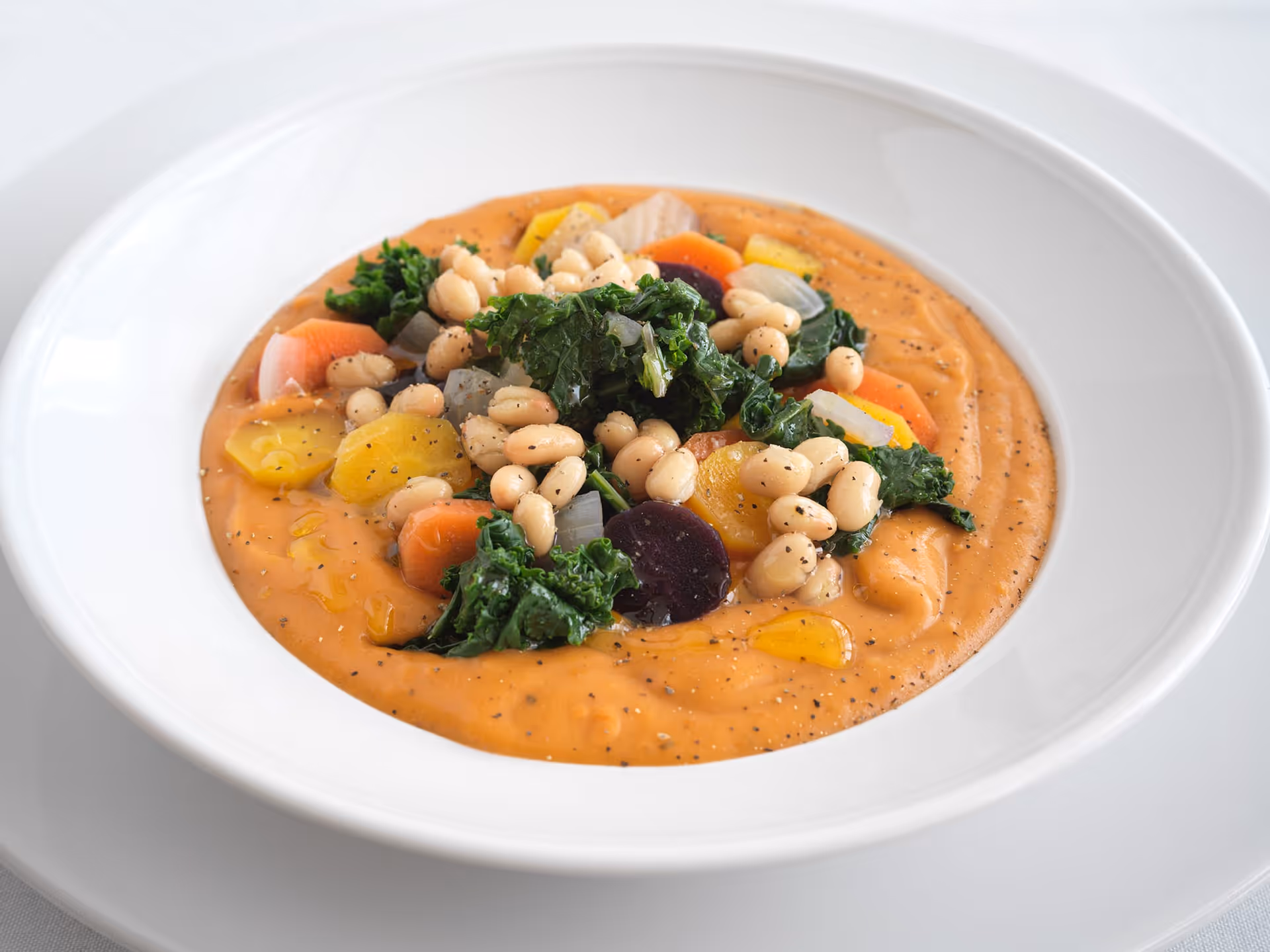 A white bowl filled with creamy orange soup topped with white beans, kale, sliced carrots, and other colorful vegetables, garnished with black pepper and a drizzle of oil.