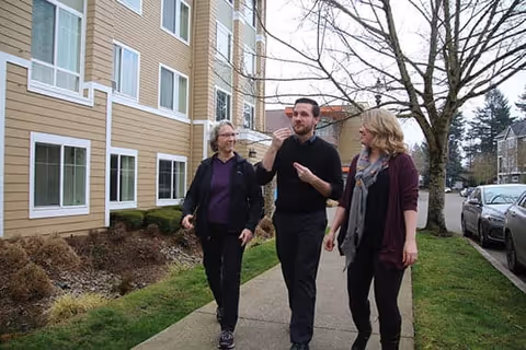 Three people walking and talking on a sidewalk next to a residential building with beige siding and white trim. There are leafless trees and parked cars along the street.