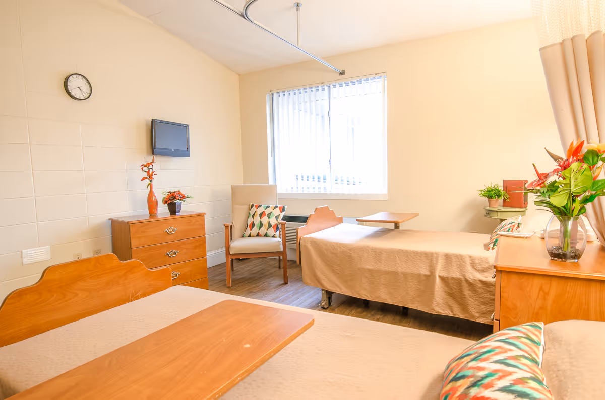 A bright and clean bedroom in a senior living facility with two single beds covered in beige bedspreads, each with a wooden overbed table. The room features a wooden dresser with a vase of flowers, a wall-mounted TV, a clock, a cushioned armchair with a patterned pillow, and a window with vertical blinds letting in natural light. There are additional plants and flowers on a side table and dresser, and beige curtains partially visible on the right side.