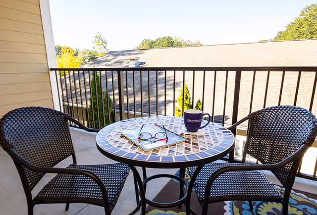 A sunny balcony with a small mosaic-top table holding a mug, reading glasses and a magazine, flanked by two wicker chairs and overlooking nearby rooftops.