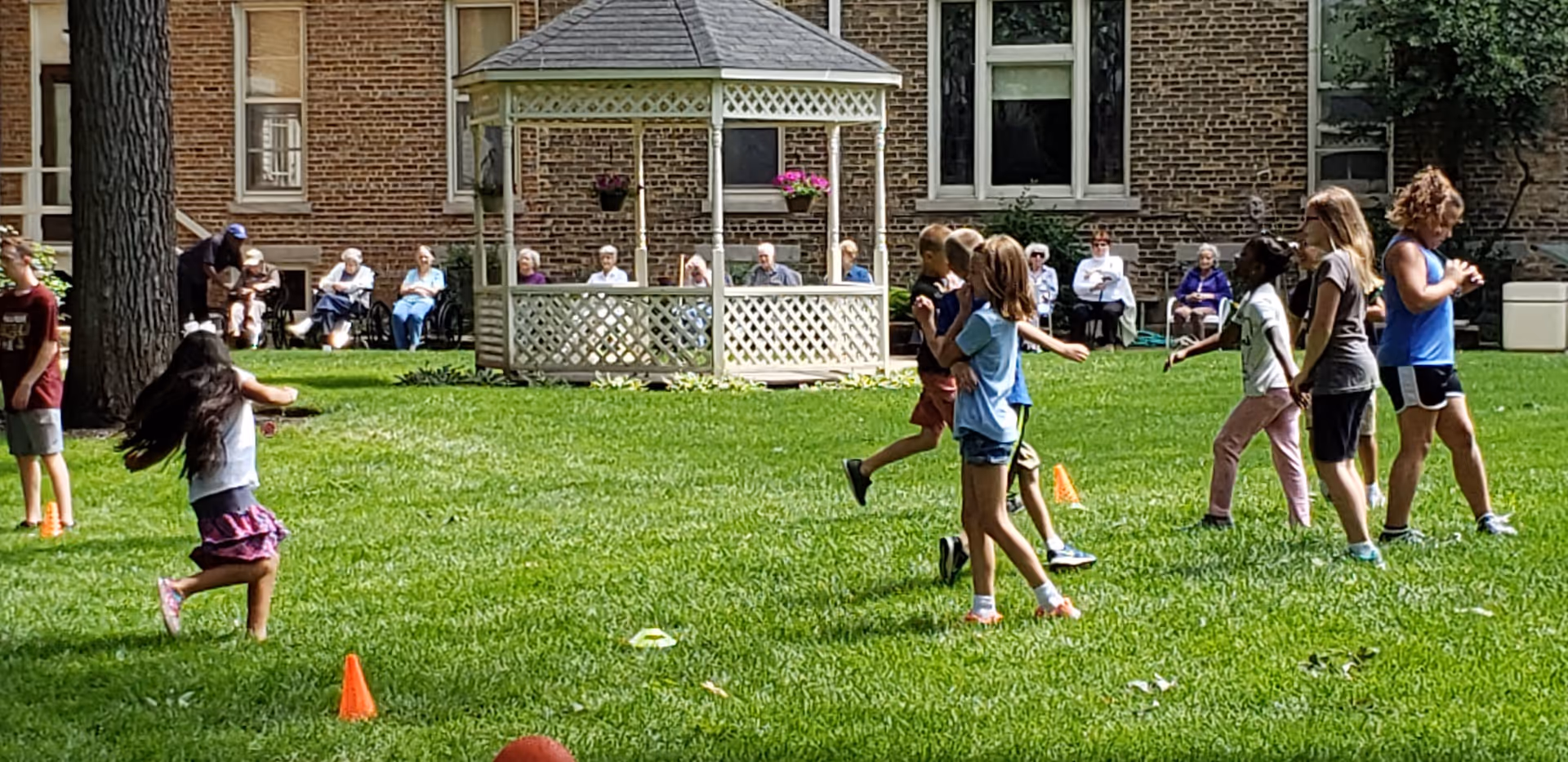 Children playing on a grassy lawn near a white gazebo with hanging flower pots, while elderly people sit in chairs along the brick building wall in the background.