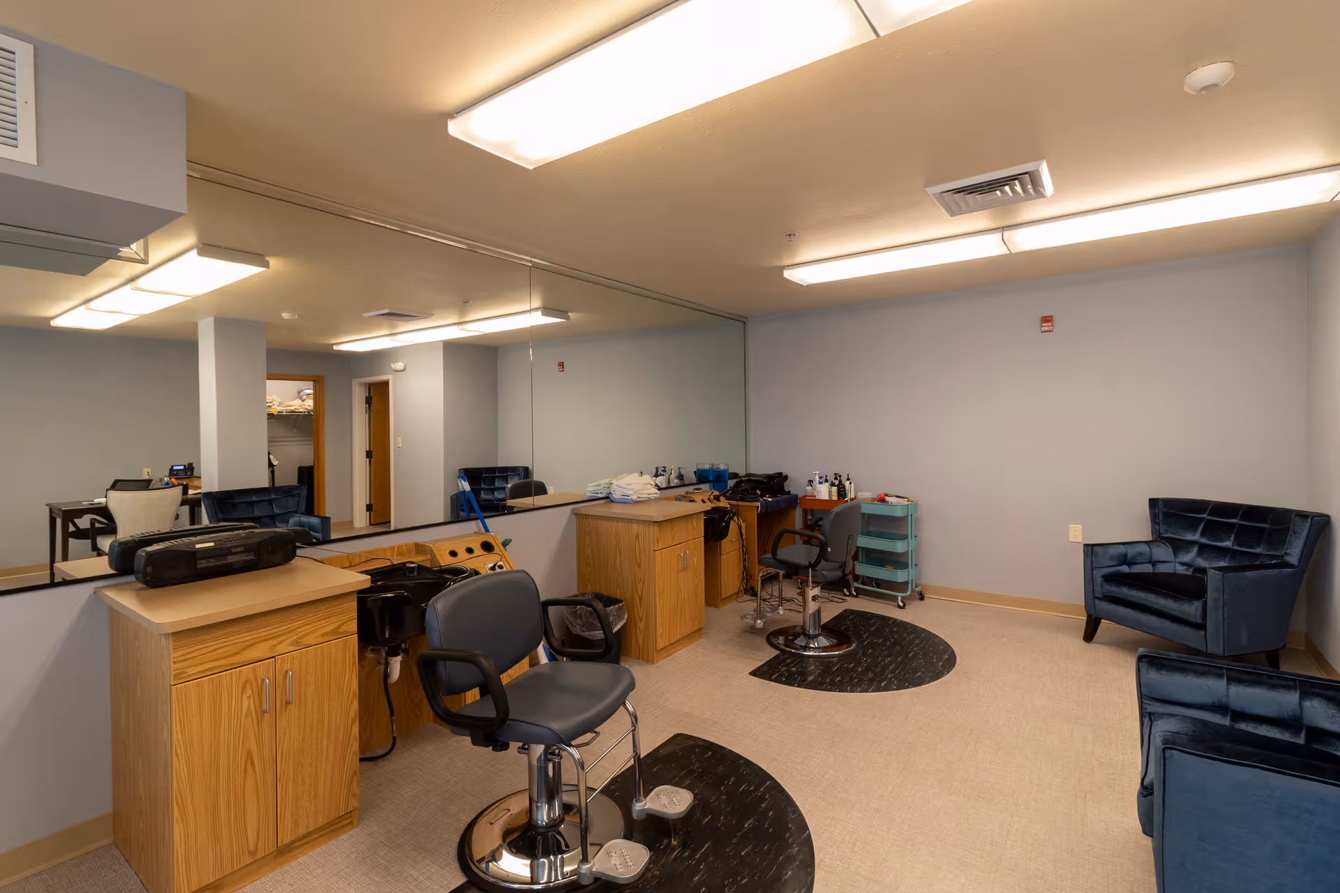 Interior of a hair salon area in a senior living facility with two black salon chairs, wooden cabinets, a large wall mirror, and two blue upholstered armchairs. The room has beige flooring and light blue walls with fluorescent ceiling lights.