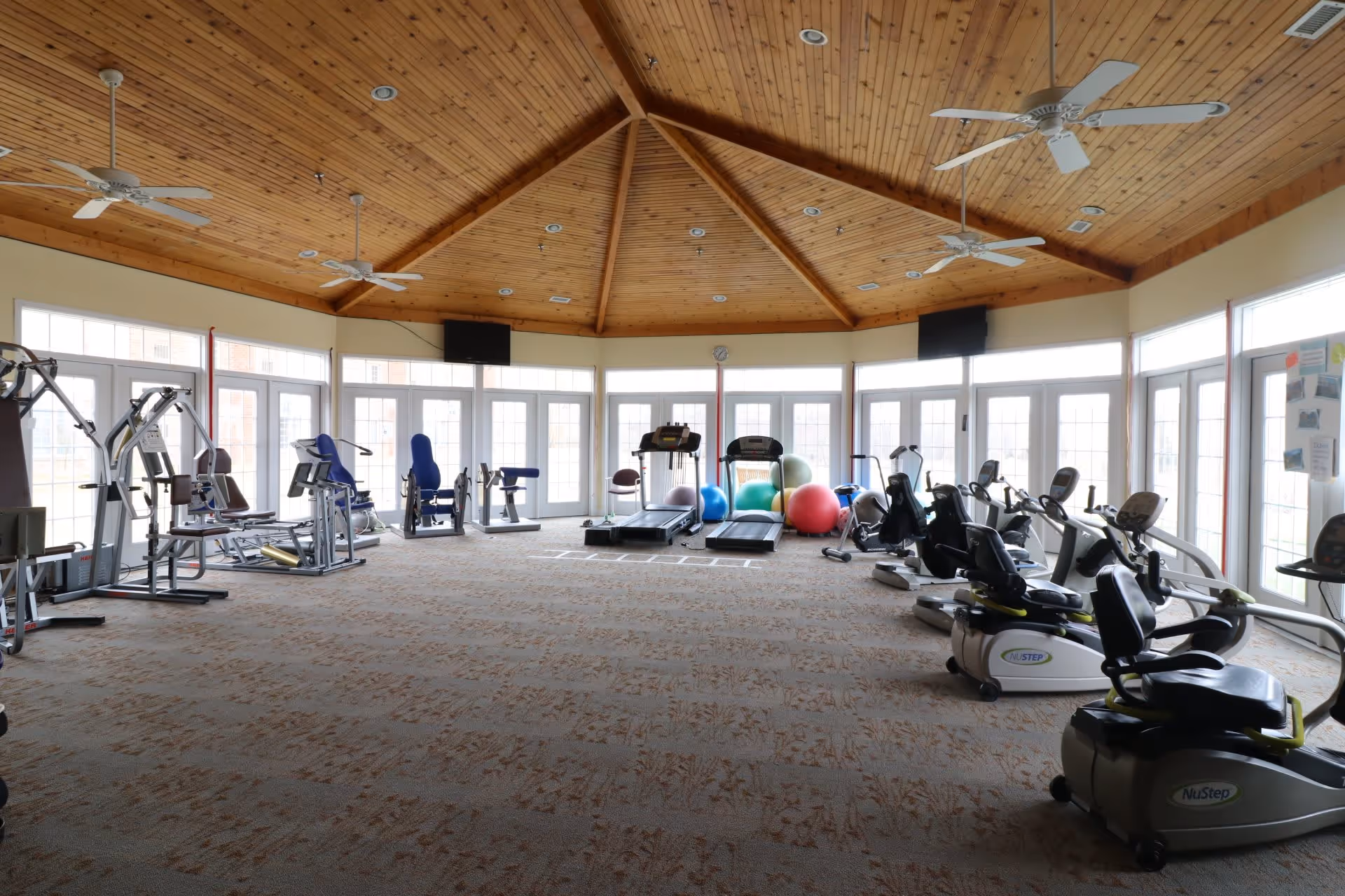 A spacious fitness room with various exercise equipment including treadmills, stationary bikes, and weight machines. The room has a high wooden ceiling with ceiling fans and large windows letting in natural light. Exercise balls are placed near the treadmills.