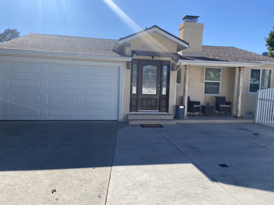 Front exterior view of a single-story house with a double garage door on the left, a central wooden front door with decorative glass panels, and a small porch area with two chairs and a small table. The house has a beige exterior with a shingled roof and two decorative columns supporting the porch roof. The driveway is concrete and spacious under a clear blue sky.