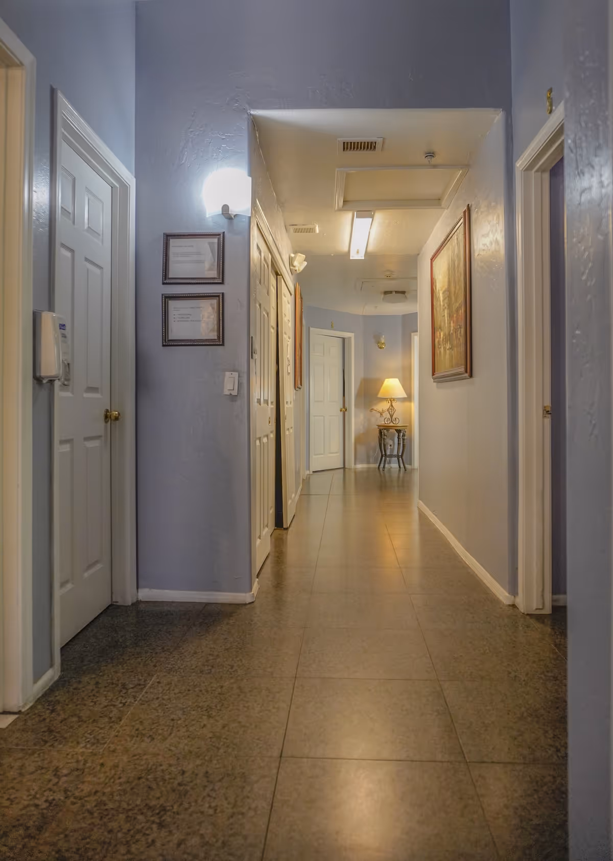 A long hallway in an assisted living home with light blue walls, multiple white doors on both sides, framed pictures on the walls, and a small table with a lamp at the far end.