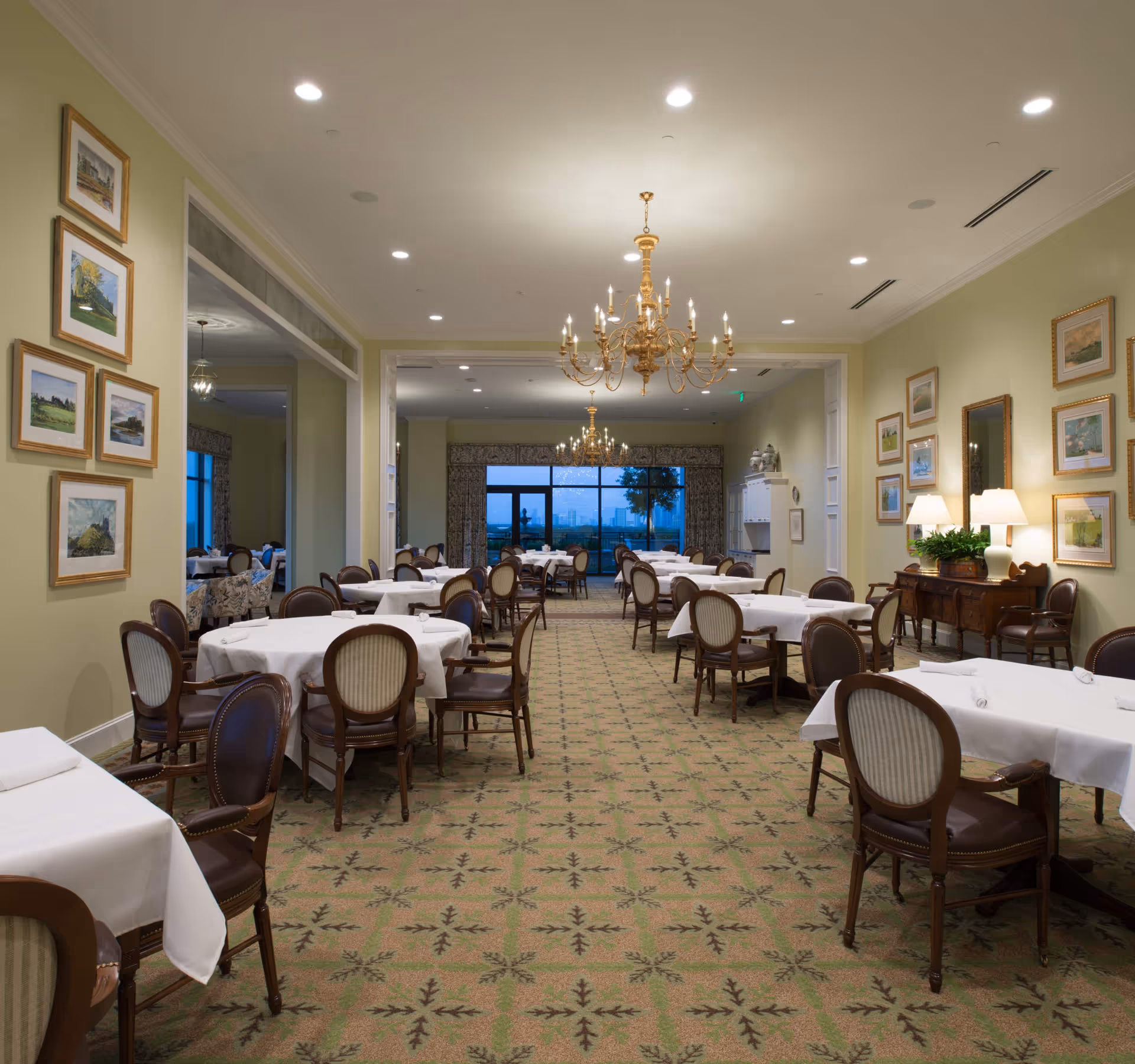 A spacious dining room with multiple round and rectangular tables covered with white tablecloths. The room features elegant wooden chairs with upholstered seats and backs, patterned carpet, framed artwork on the walls, and two ornate chandeliers hanging from the ceiling. Large windows at the far end provide a view outside.
