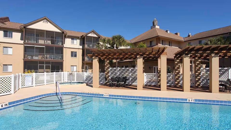 Outdoor swimming pool with clear blue water in front of a multi-story residential building. The pool area includes a hot tub, a white lattice fence, and shaded seating areas under wooden pergolas. Palm trees and clear blue sky are visible in the background.