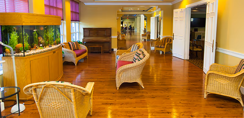 Bright communal sitting area with wicker chairs, a fish tank and a piano on polished hardwood floors.