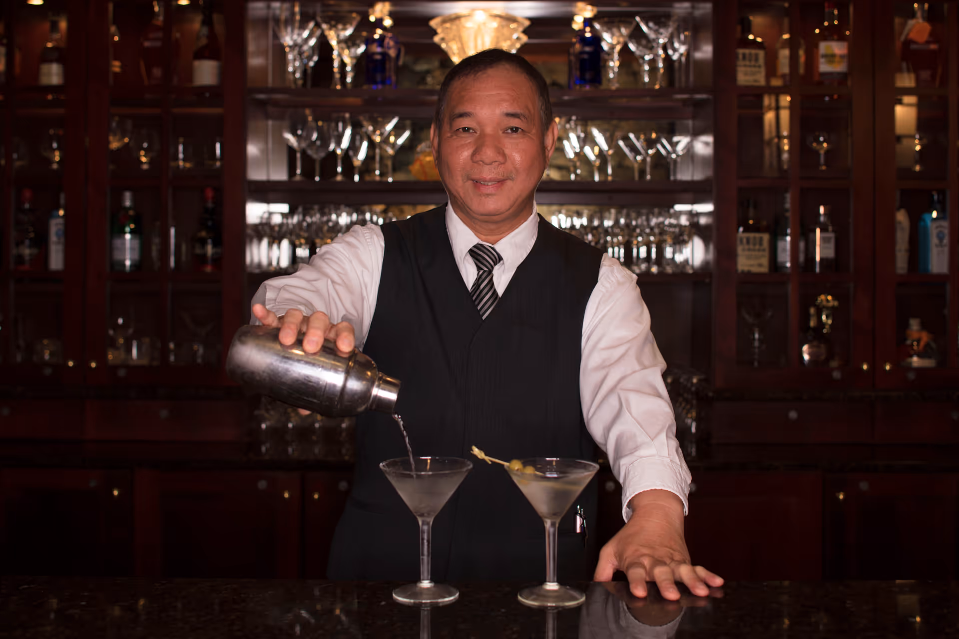 A bartender wearing a white shirt, black vest, and striped tie is pouring a drink from a cocktail shaker into a martini glass on a bar counter. Another martini glass with olives on a toothpick is placed next to it. Behind him is a wooden cabinet filled with various bottles and glassware.