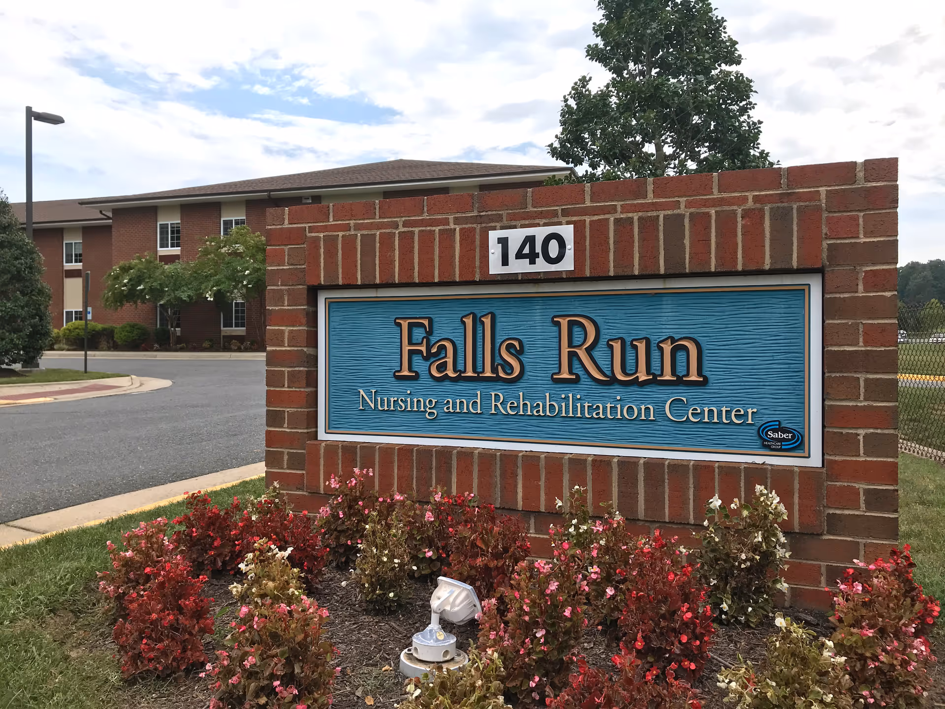 A brick sign for Falls Run Nursing and Rehabilitation Center with the number 140 above it. The sign is surrounded by a flower bed with red and pink flowers. In the background, there is a multi-story brick building, a tree, a street lamp, and a partly cloudy sky.